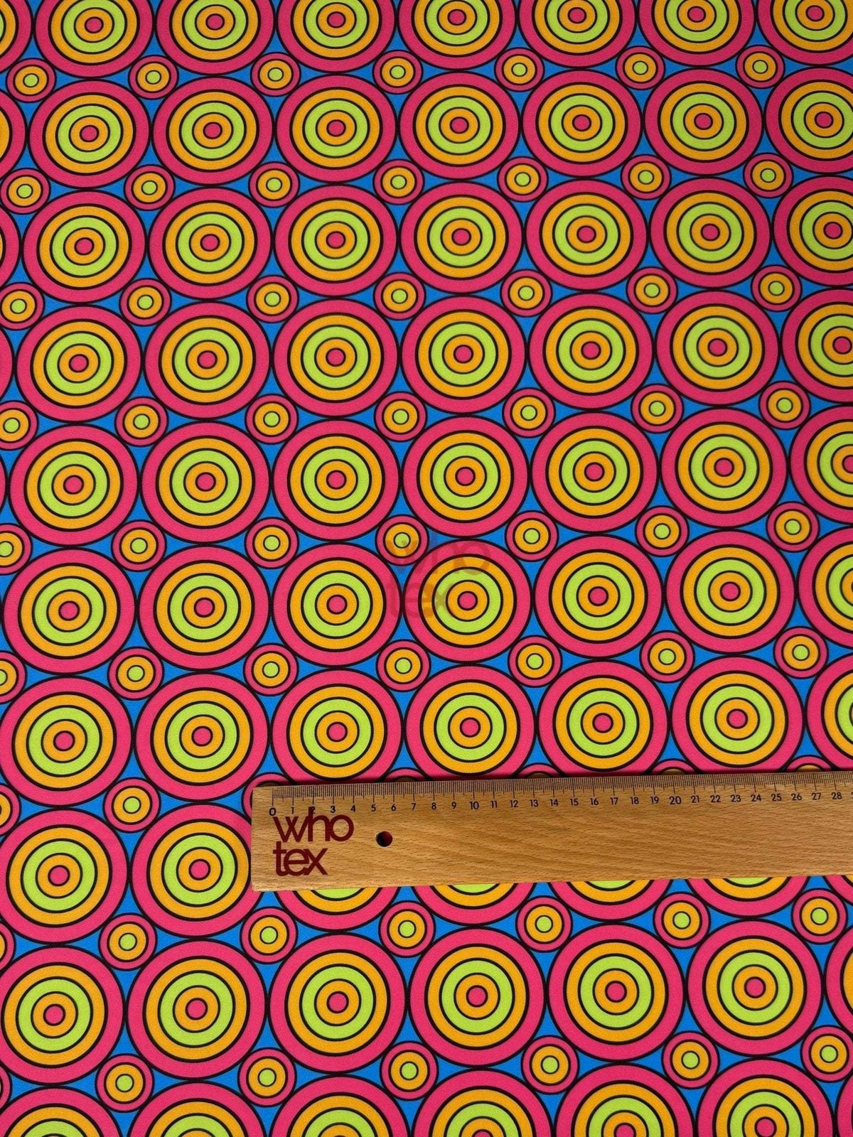 Colorful geometric circles print fabric with vibrant concentric circles in red, yellow, green, and blue, shown with a wooden ruler for scale