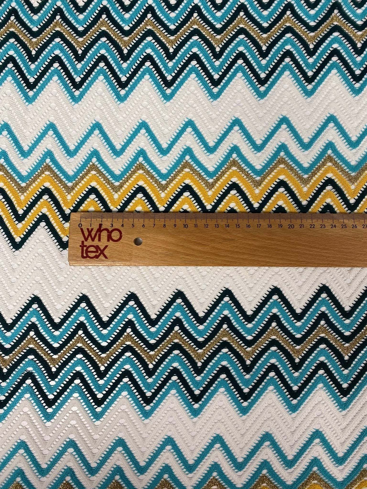 close-up of geometric zigzag embroidered fabric with green, teal, white, black, and mustard yellow pattern and a wooden ruler for scale
