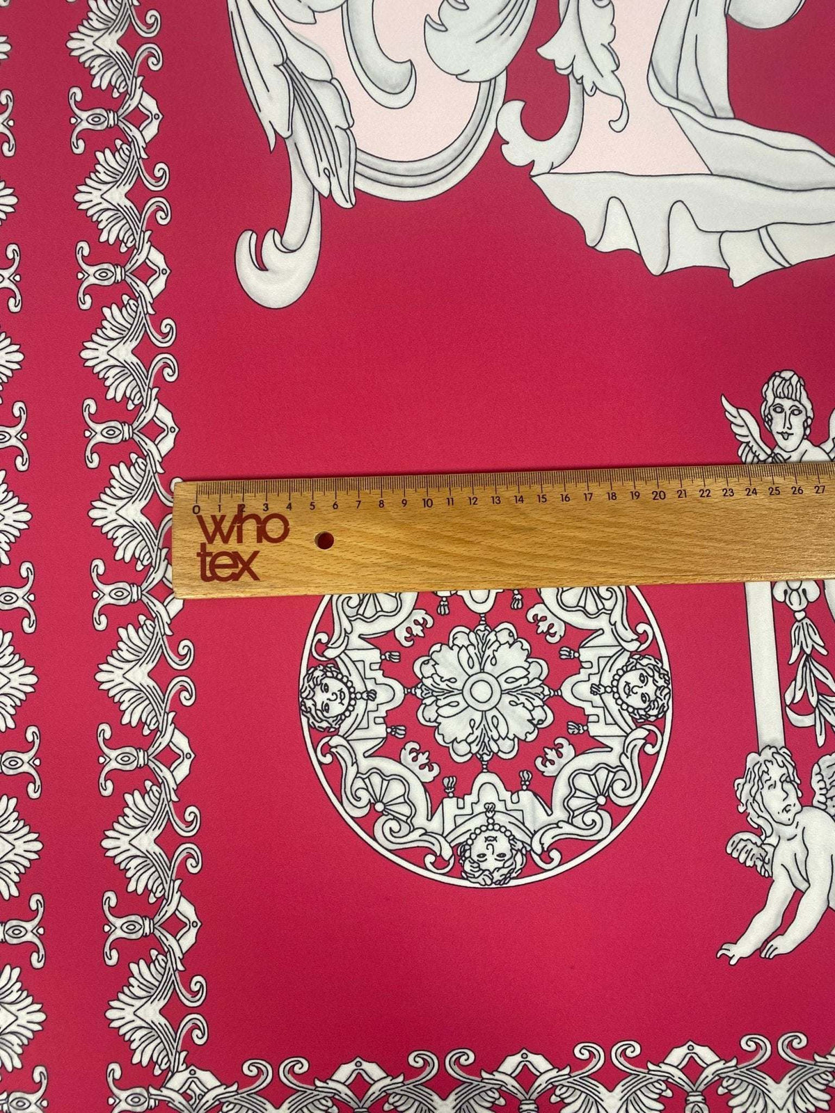 Pink vintage Baroque fabric with intricate cherub and ornate pattern next to a wooden ruler showing scale